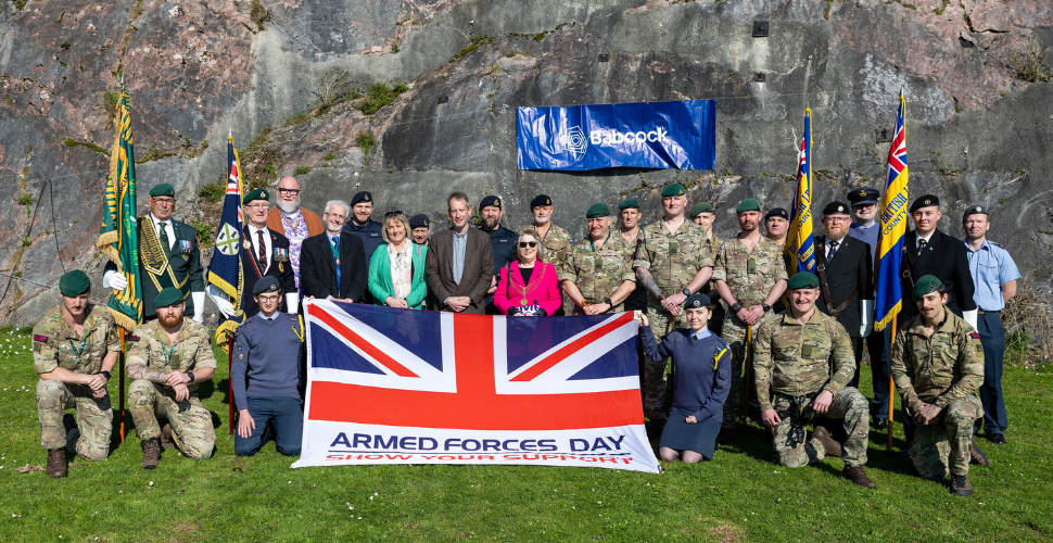 Group of armed forces personnel and civilians posing outdoors on grass, holding an Armed Forces Day Union Jack banner with flags and a Babcock sign displayed behind them against a rocky backdrop.
